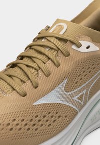 Μπεζ αθλητικό παπούτσι με επάνω μέρος από δίχτυ, μπεζ κορδόνια, λευκό λογότυπο στο πλάι και μαλακή λευκή σόλα σχεδιασμένη για τρέξιμο ή αθλητισμό.