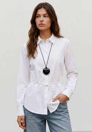 Femme aux longs cheveux bruns portant une chemise blanche boutonnée avec une taille nouée et un jean bleu, accessoirisée d'un collier pendentif rond noir.
