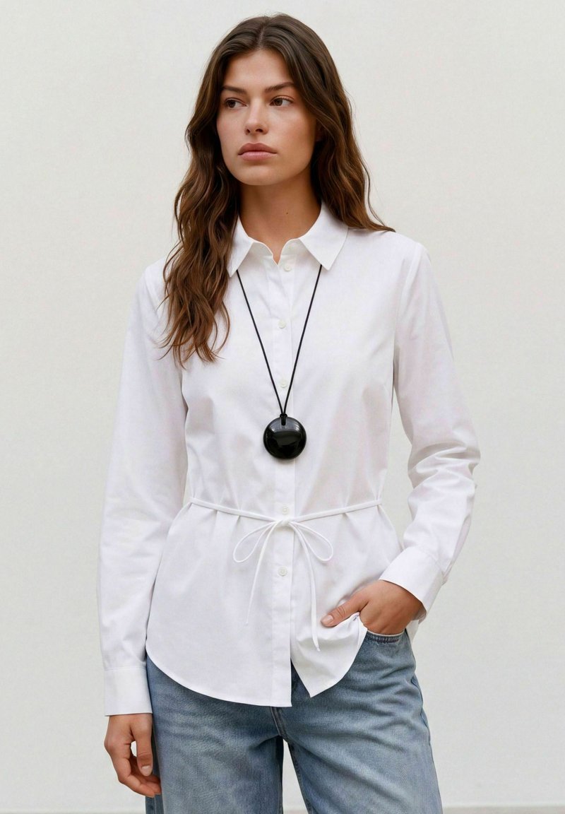 Femme aux longs cheveux bruns portant une chemise blanche boutonnée avec une taille nouée et un jean bleu, accessoirisée d'un collier pendentif rond noir.
