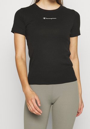 Femme portant un t-shirt noir ajusté Champion à manches courtes et des leggings beige sur un fond uni.