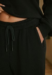 Schwarze, strukturierte Jogginghosen mit elastischem Bund und Kordelzug. Seitentaschen sind sichtbar; der Stoff wirkt weich und leicht.