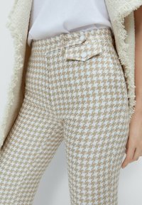 Beżowe spodnie w wzór houndstooth w kolorze białym z przodu z guzikami, w wysokim pasie i teksturowanym materiale, w zestawie z beżową kurtką ozdobioną frędzlami.