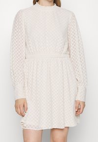 Vestido largo de manga larga en un tono beige claro, con un patrón geométrico texturizado, cintura fruncida y un escote alto con volantes. Tela suave y fluida.