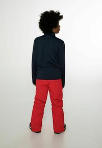 Una persona vista da dietro indossa una maglia blu navy a maniche lunghe e dei pantaloni da neve rossi. La maglia ha un colletto alto; i pantaloni hanno una vestibilità comoda.