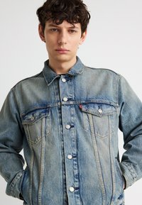 Νεαρός άνδρας με σγουρά μαλλιά που φοράει ανοιχτό μπλε τζιν μπουφάν με ασημένια κουμπιά, τα χέρια στις τσέπες, στέκεται μπροστά σε απλό λευκό φόντο.