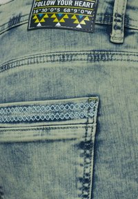 Jeans in denim azzurro chiaro con una piccola tasca frontale, cuciture decorative in azzurro chiaro e una patch con il testo "SEGUITA IL TUO CUORE".