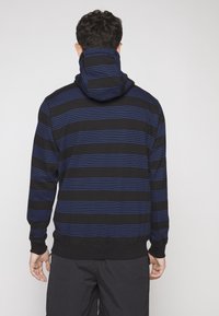 Felpa nera con strisce orizzontali blu, dotata di cappuccio e polsini a costine. Texture morbida e design casual, adatta per un look informale.