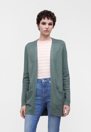 Jeune personne aux cheveux courts et bouclés porte un cardigan vert ouvert, une chemise rose clair rayée et un jean bleu, debout devant un fond uni.