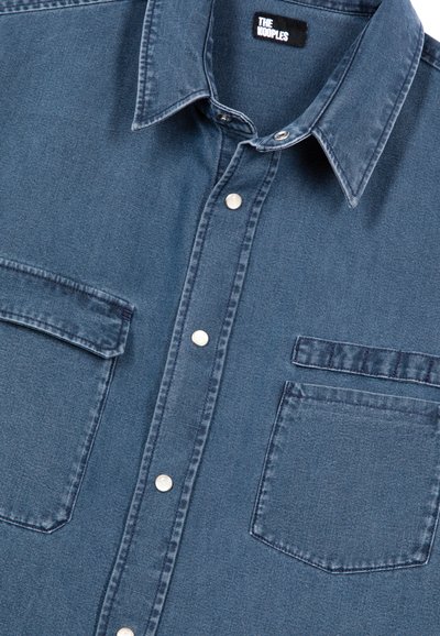 Camicia in denim blu scuro con colletto a punta, due tasche frontali, bottoni argentati e un'etichetta di marca visibile, caratterizzata da un tessuto con texture.