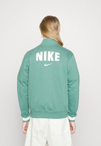 Grön sweatshirt i strukturerad tyg med vit "NIKE"-text på baksidan och en liten Nike swoosh-logotyp, med ribbade ärmslut.