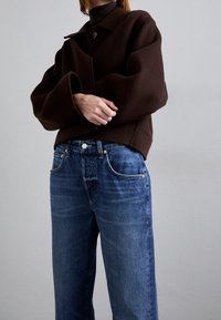 Brun oversized jacka med krage och knapprad, kombinerad med blå raka jeans med koppardetaljer och en använd känsla.