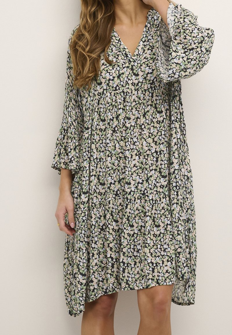 Femme portant une robe ample longueur genou avec un petit motif floral noir, blanc et vert et des manches cloches trois-quarts.