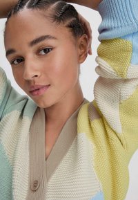 Neulekahvijaki, jossa on sekoitus pastellivärejä, v-mallinen kaula-aukko, teksturoitu kuvio ja yksi nappikiinnitys.