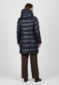 Námořnický pufovaný kabát s lesklým povrchem, který má kapuci, horizontální prošívání a délku ke kolenům. V kombinaci s hnědými širokými kalhotami.