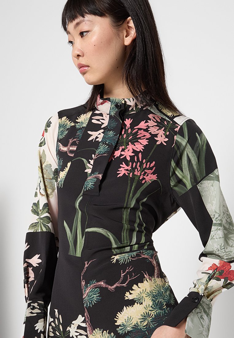 Zwarte bloempatron blouse met lange mouwen, een hoge kraag en een knopdetail bij de hals. Verschillende levendige plantontwerpen overal.