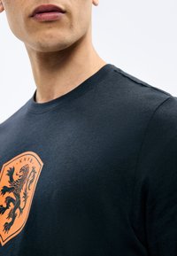 Κλείσιμο σε πρόσωπο που φοράει σκούρο ναυτικό πουκάμισο με πορτοκαλί έμβλημα σε σχήμα ασπίδας που φέρει μαύρο λιοντάρι και το κείμενο "KNYB".