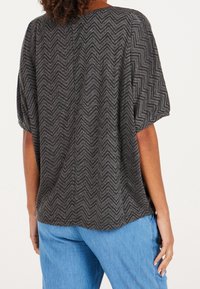 Osoba nosící volné černé a šedé tričko s krátkými rukávy a cikcak vzorem, v kombinaci se světle modrými džíny, zobrazená zezadu.