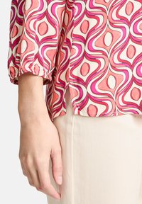 Bunte Bluse mit geometrischem Muster in Pink-, Rot- und Weißtönen. Leichter Stoff, lockere Passform und geraffte Bündchen.