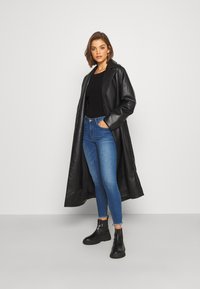 Palton din piele neagră cu rever larg, asortat cu un pulover texturat negru și blugi ajustați albaștri; modelul poartă cizme negre până la gleznă.