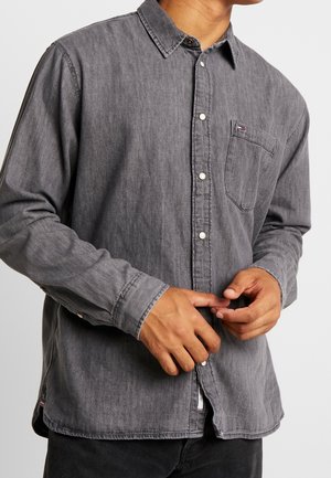 Homme portant une chemise boutonnée gris foncé délavée avec une poche poitrine et des boutons blancs visibles, ajustant les poignets.