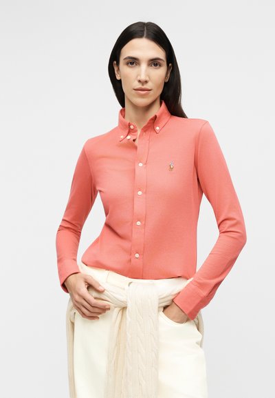 Pikkade tumepruunide juustega naine, kes kannab korallivärvi nööpidega särki ja kreemika värvusega pükse ning tal on vöökohal seotud kreemja värvusega kudumiskampsun.