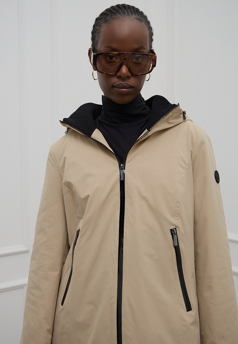 Beige waterdichte jas met een zwarte capuchon, voorrits en twee zijzakken. De stof heeft een soepele textuur en bevat zwarte accenten.