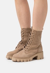 Botas de tobillo color marrón con cordones, hechas de ante, con una suela de goma gruesa y costuras detalladas. El diseño es resistente y tiene un acabado suave.