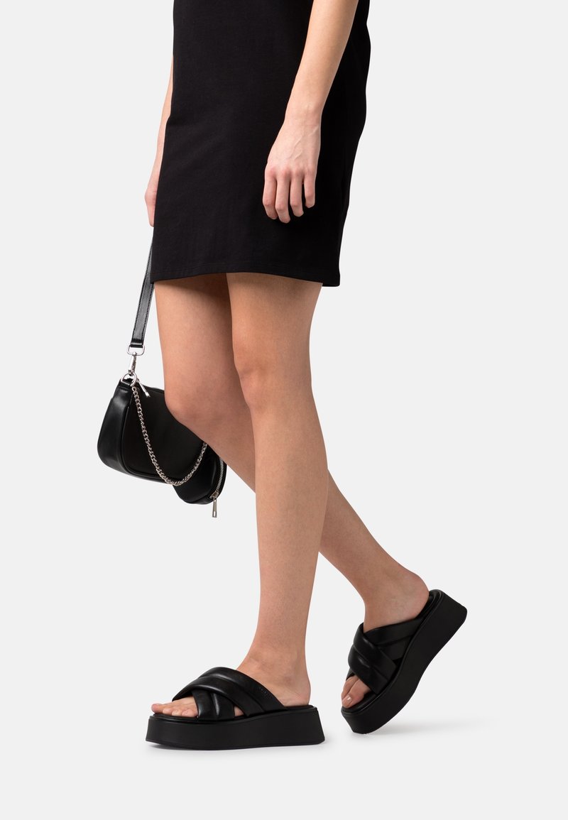 Femme portant une robe courte noire et des sandales plates-formes noires, tenant un petit sac à main noir avec une bandoulière chaîne.