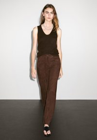Pantaloni scamosciati marrone scuro con taglio dritto, abbinati a un top senza maniche nero e sandali neri. Accento di una semplice collana in argento.