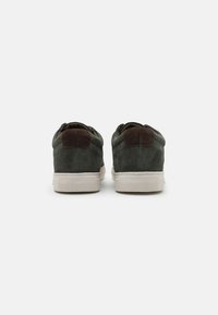 Zapatos de ante verde oscuro con acentos de ante marrón en el tacón y suelas de goma blancas, que presentan una superficie texturizada y un diseño minimalista.