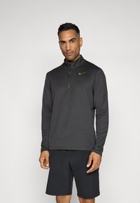 Crna duga rukava sportska majica s polu-rajseflerom i teksturiranim materijalom. Ima kontrastni zlatni Nike logo na prsima. Uparena s crnim šortsevima.