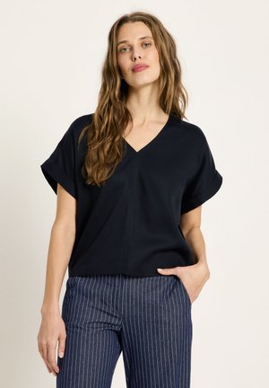 Woman with long wavy hair wearing a black V-neck short-sleeve top and navy pinstripe pants, standing with one hand in pocket.