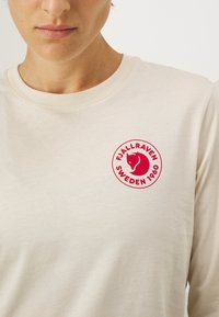 Bež pika vormis T-särk pikkade varrukatega, ümmarguse kaelusega ja punase ringikujulise logoga, kus on kujutatud rebase siluetti ja tekst: "FJÄLLRÄVEN SWEDEN 1960."