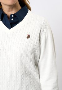 Hvit kabelstrikket genser med v-hals. Har et teksturert mønster og en liten brodert logoakcent på brystet.
