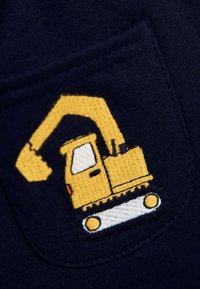 Tumši zils audums ar dzeltenu izšūtu ekskavatora dizainu, kurā redzama detalizēta kabīne, roka un kājas. Novietots uz kabatas.