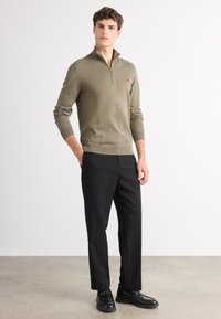 Pullover lavorato a maglia verde oliva con zip a un quarto, design attillato, polsini e orlo a coste; abbinato a pantaloni neri e scarpe nere.