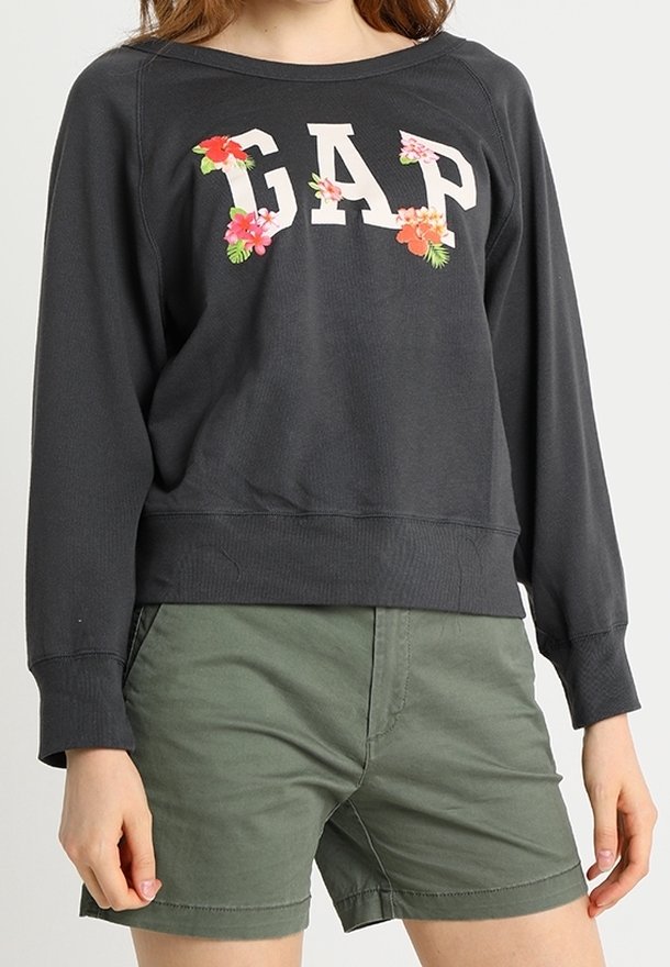 Personne portant un sweat-shirt GAP noir à manches longues avec des accents floraux, associé à un short vert, debout, les bras relâchés le long du corps.