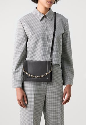 Black leather crossbody bag with a gold chain accent, featuring a rectangular shape and smooth surface. Worn with a checkered patterned outfit.