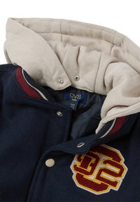 Chaqueta de forro polar azul marino con forro de capucha beige, cuello a rayas rojas y blancas, y parche con número bordado. Se cierra con broches.