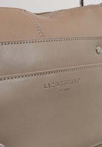 Beige läderhandväska med slät textur, sydda detaljer och pr präglad logotyp. Har ett dragkedjestängning och silverfärgad hårdvara.