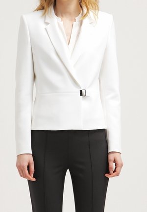 Femme portant un blazer blanc ajusté avec un détail de boucle à l'avant et un pantalon noir sur mesure sur un fond uni.