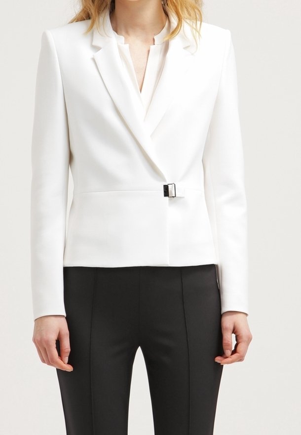 Femme portant un blazer blanc ajusté avec un détail de boucle à l'avant et un pantalon noir sur mesure sur un fond uni.