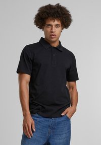 Svart polo skjorte laget av bomull, med krage, korte ermer og en placket med tre knapper; brukt med blå denimjeans.