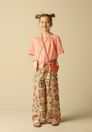 Fille souriante avec double chignons porte un t-shirt pêche, un pantalon à jambes larges à motifs, deux sacs bandoulière perlés et des sandales sur fond beige.