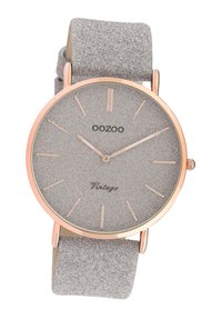 Montre ronde métallique avec un cadran gris texturé, des accents en or rose et un bracelet doux gris. Marqueurs d'heures minimalistes et nom de la marque affiché.