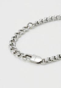 Pulsera de cadena de metal plateado con eslabones entrelazados y un cierre. La superficie es brillante con una textura suave e incluye un detalle de marca.