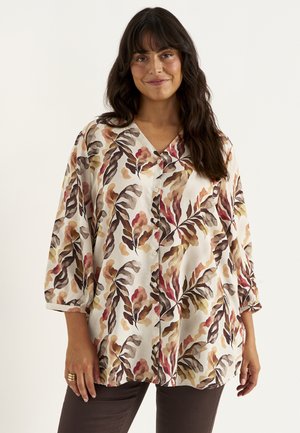 Vrouw met donker haar draagt een losse blouse met knopen en een meerkleurig bladpatroon en een donkere broek, staand tegen een effen achtergrond.