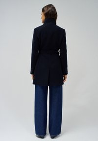 Manteau en laine noir avec un col haut, une ceinture à la taille et des boutons aux poignets. Associé à un pantalon en jean bleu à jambes larges.