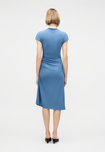 Γυναίκα με κοντά ξανθά μαλλιά, ντυμένη με εφαρμοστό μπλε midi φόρεμα και μαύρα mule με τακούνι, στέκεται με την πλάτη γυρισμένη προς μία απλή λευκή επιφάνεια.