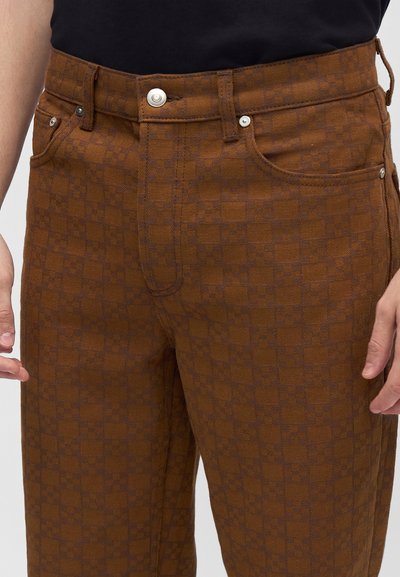 Pantalon marron à carreaux avec fermeture par bouton argenté porté avec un haut noir, montrant des poches avant et des coutures détaillées sur le tissu.